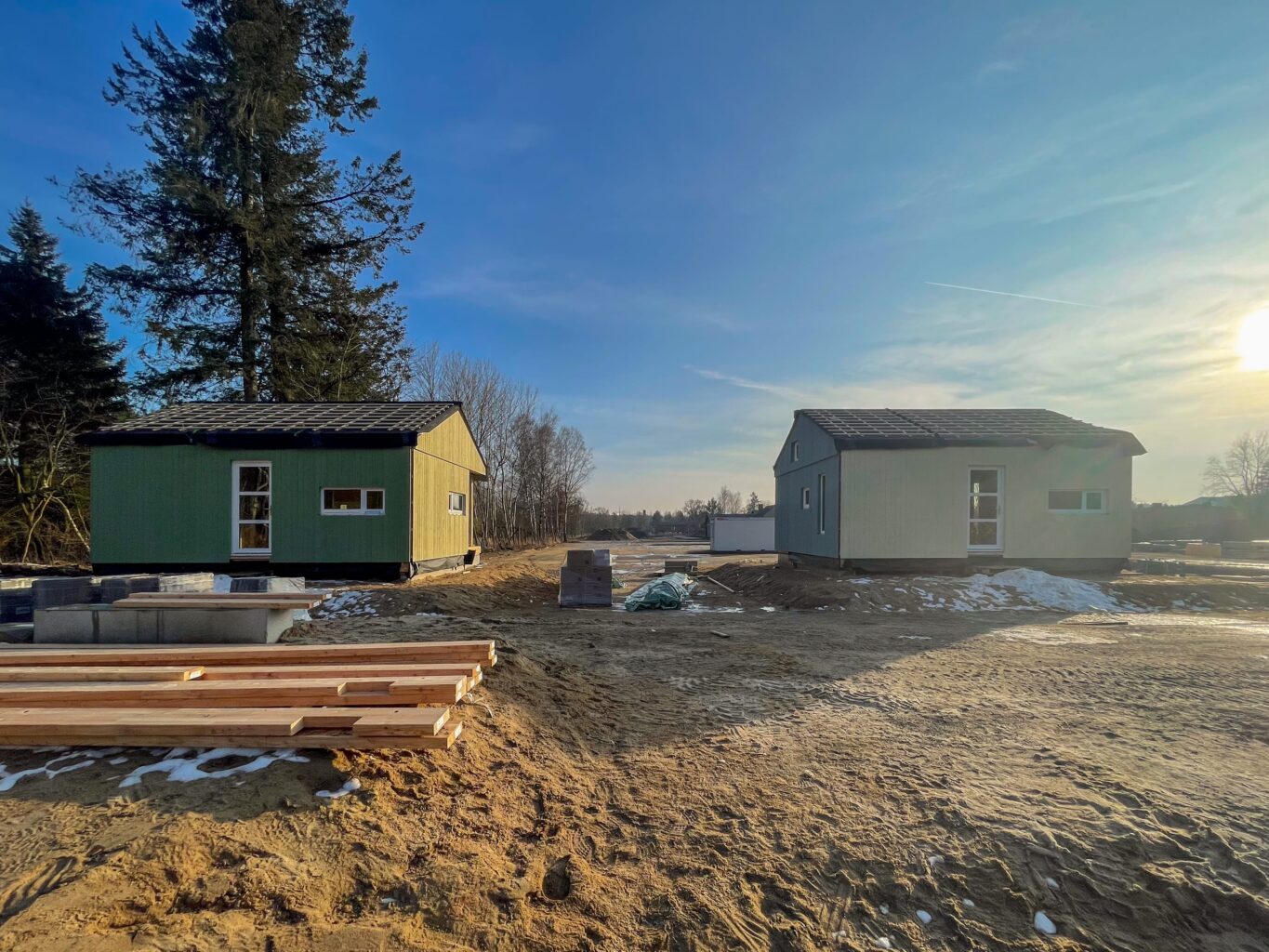 Zwei kleine Holzhäuser im Bau, eines grün und eines grau, stehen auf sandigem Boden mit Baumaterialien und Schneeresten bei tiefstehender Sonne.