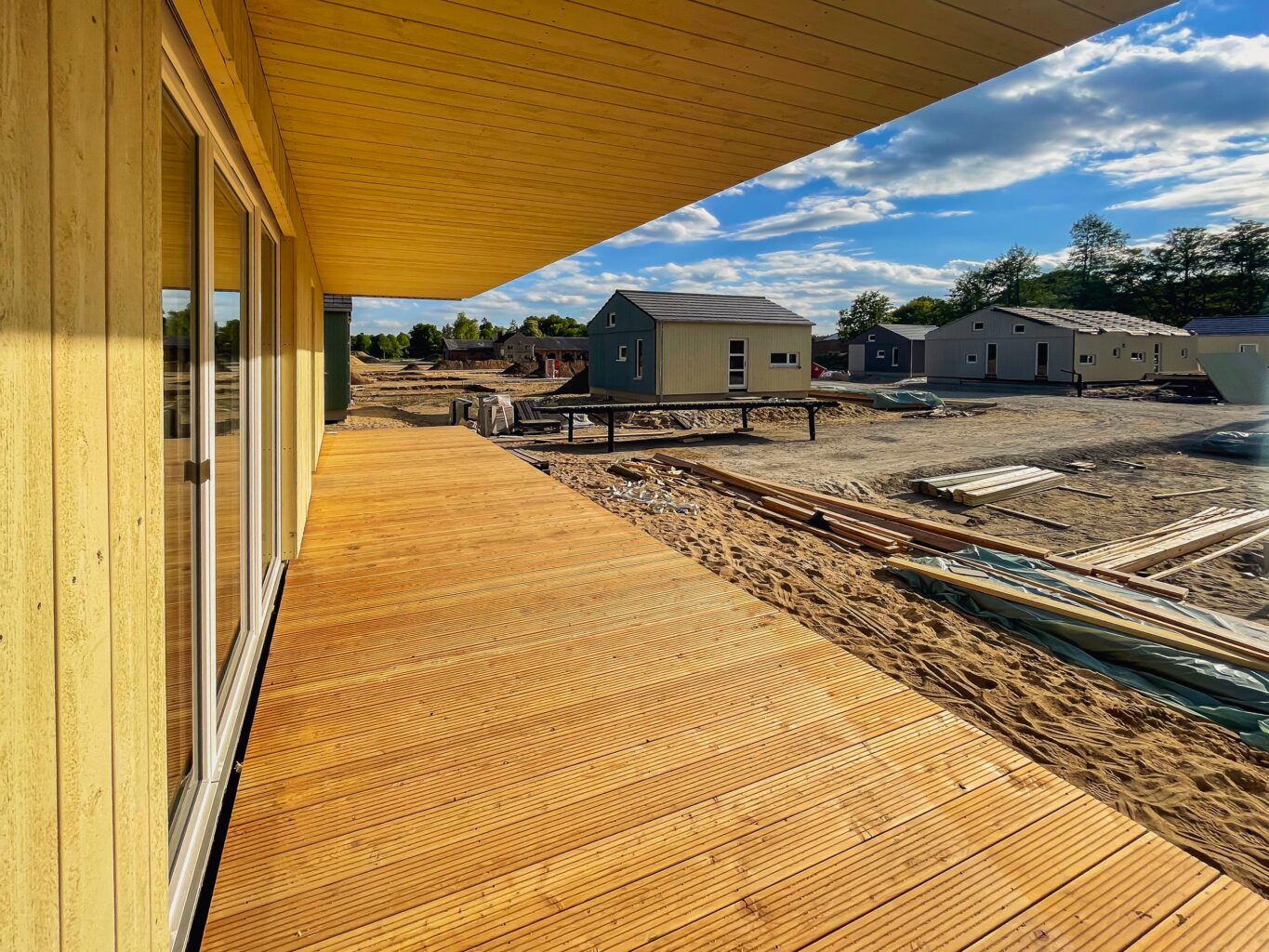 Blick auf eine neue Holzterrasse mit Dachüberstand, umgeben von mehreren im Bau befindlichen Holzhäusern unter blauem Himmel.